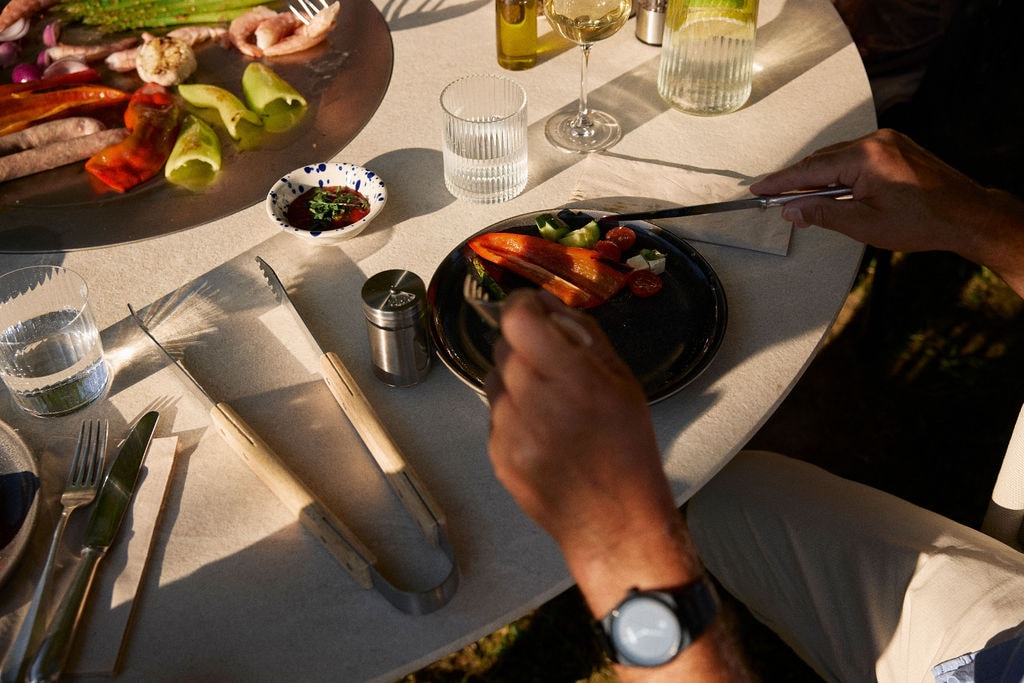 Menschen sitzen um einen runden Grilltisch, kochen gemeinsam und genießen die Atmosphäre im Freien.